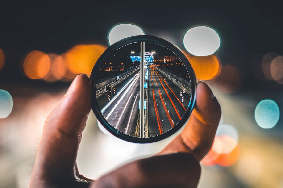 Hand holding a lens showing a sharp corrected image of a highway scene, symbolizing multi-sensor fusion and depth imaging capabilities at Ability Enterprise
